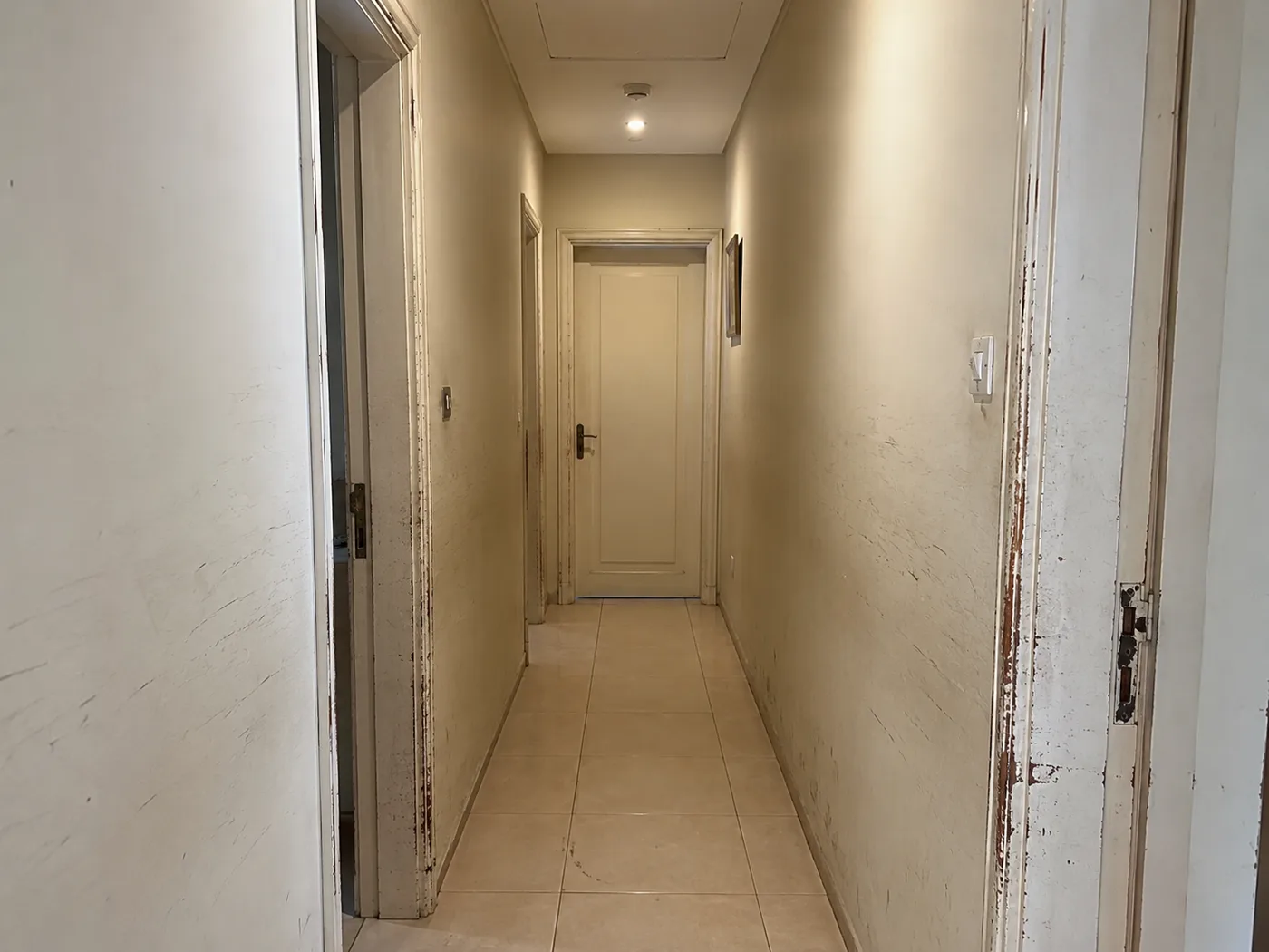 A narrow hallway with scuffed cream walls and old white door frames chipped back to bare wood